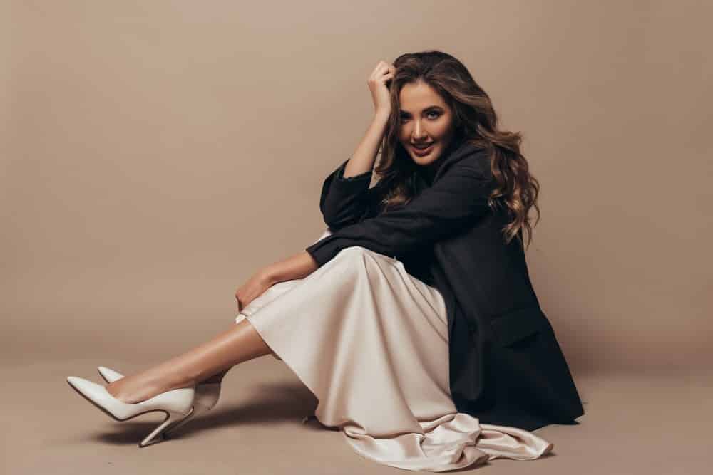 cheerful-model-sitting-floor-wearing-modern-oversize-black-jacket-creamy-long-dress-high-heel-shoes-her-feet-curly-hairstyle-makeup (1)