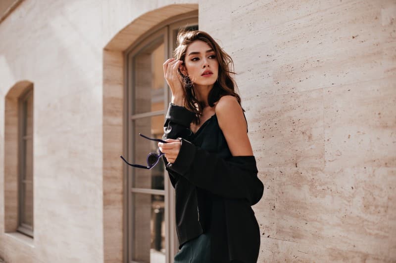 cute-young-girl-with-dark-wavy-hairstyle-bright-makeup-silk-dress-black-jacket-holding-sunglasses-hands-looking-away-against-beige-building-wall (1)